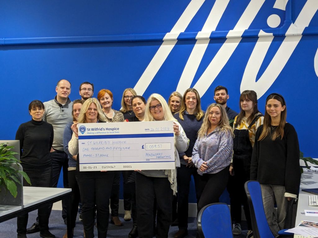 MindWorks Marketing colleagues presenting the charity cheque to St Wilfrid's Community & Corporate Fundraiser, Vanessa. They are standing in front of a blue wall painted with MindWorks' logo.