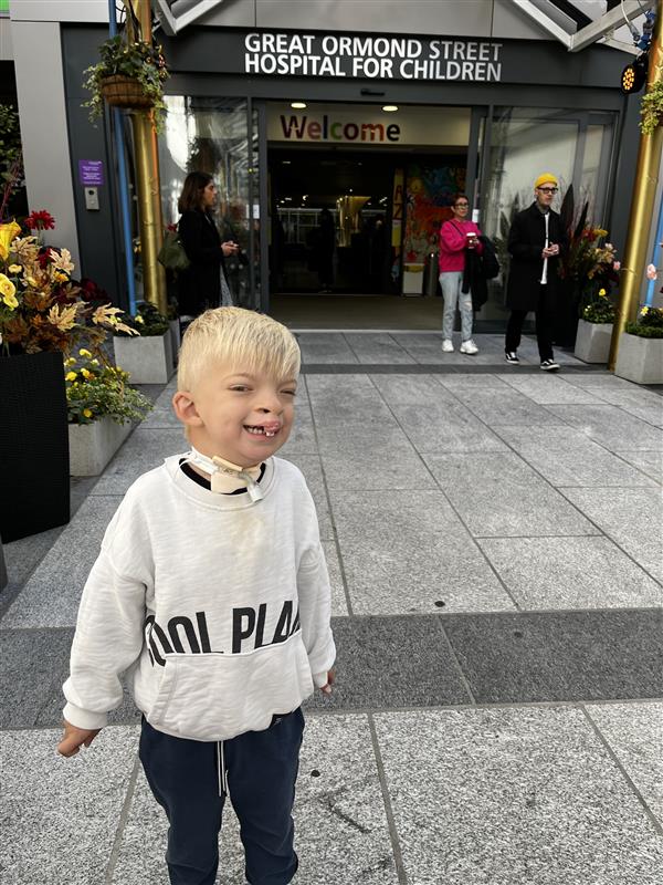Dougie, a young, blond boy with a tracheostomy stands smiling happily in front of the entrance to Great Ormond Street Hospital. He's wearing a grey sweatshirt and black jogging bottom-type trousers.