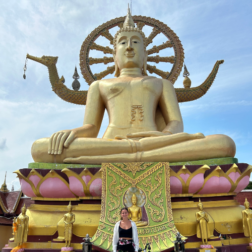 Melissa stands in front of a large golden Buddha statue in a seated position in South East Asia. The statue features intricate details, including a halo-like structure behind the Buddha's head and two ornate dragon-like extensions on either side. The base of the statue is decorated with smaller golden statues and a richly patterned green and gold cloth. The sky is clear with a few clouds, providing a bright backdrop to the impressive structure. Melissa is wearing a black top with floral patterns and has her hair pulled back, standing at the center bottom of the image, directly in front of the statue.