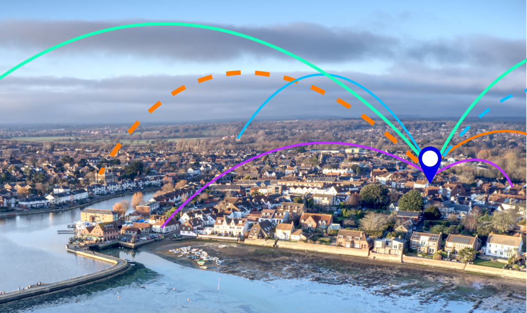 Aerial view of coastal town, Emsworth, with a blue location pin denoting MindWorks Marketing. Colorful, curved lines in green, blue, orange, and purple emanate from the pin, symbolizing its connection to the city of Portsmouth and the South. The town features a mix of houses and buildings near the water, with a small harbor in the foreground and a cloudy sky overhead.