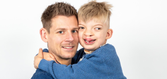 A picture of Adam holding his son Dougie. Dougie has his arms wrapped around Adam's neck. Their heads are touching as they both look to the camera. Set on a white studio background, they are both wearing blue jumpers.