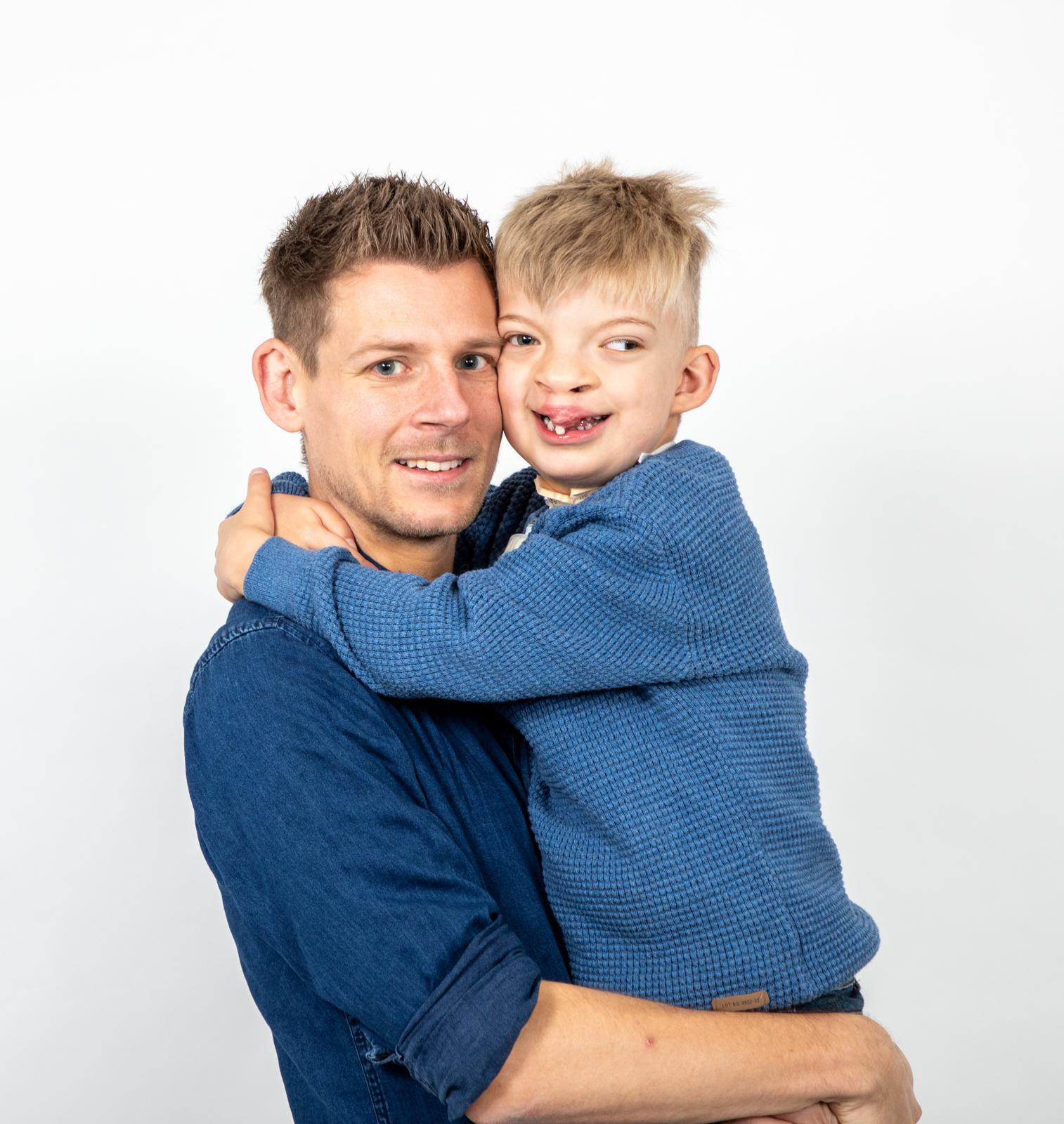 A picture of Adam holding his son Dougie. Dougie has his arms wrapped around Adam's neck. Their heads are touching as they both look to the camera. Set on a white studio background, they are both wearing blue jumpers.